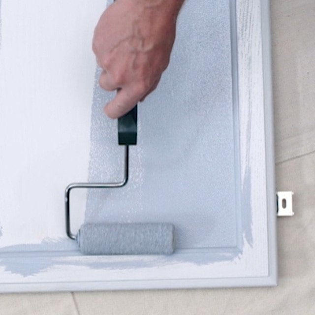 A person rolling primer onto a white painted kitchen cabinet door that is lying flat on a white drop cloth. A person rolling primer onto a white painted kitchen cabinet door that is lying flat on a white drop cloth.